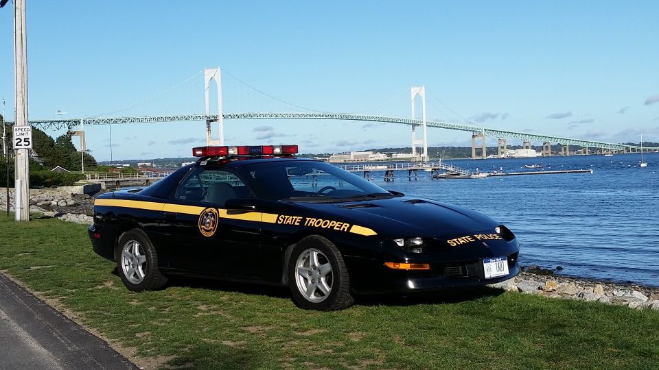 Black state trooper car with yellow stripe and "STATE TROOPER" text, parked by the water with a bridge in the background. License plate reads "111 787"