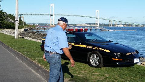 1991 Ford LTD Crown Victoria - A black state trooper car is parked near the water with a bridge in the distance under a clear blue sky.