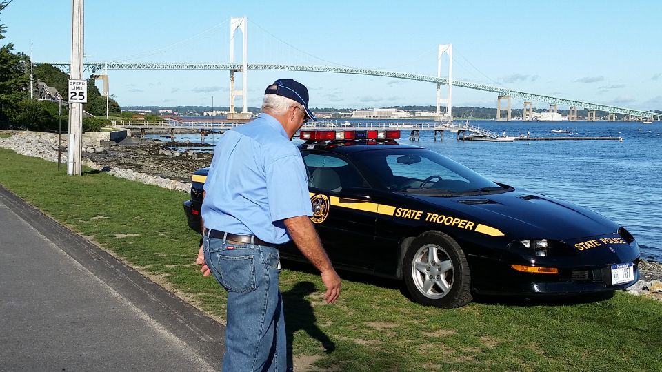 State trooper car parked by water with a bridge in the background. Man in blue shirt stands nearby