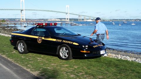 1991 Ford LTD Crown Victoria - A black police car with a yellow stripe and "STATE TROOPER" text is parked by the water with a bridge in the background.