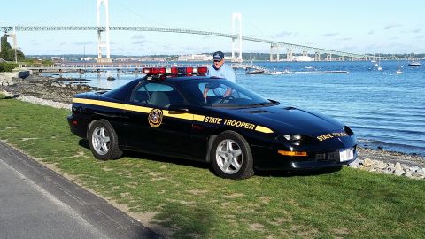 1991 Ford LTD Crown Victoria - A black police car, marked "STATE TROOPER" with a yellow stripe, is parked near the water with a bridge visible in the distance.
