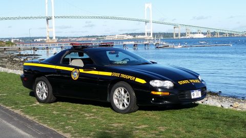 1991 Ford LTD Crown Victoria - A black state trooper car is parked on grass near the water, with a large bridge visible in the distance.