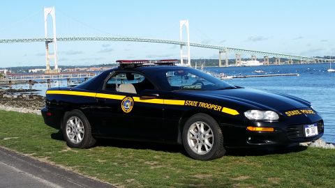 1991 Ford LTD Crown Victoria - A black state trooper car is parked on a grassy area next to the water, with a large bridge visible in the distance.