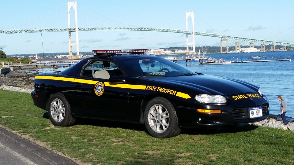 Black state trooper car with yellow stripe and "STATE TROOPER" text parked by water with a bridge in background. License plate reads 3567