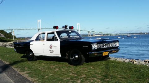 1991 Ford LTD Crown Victoria - A vintage police car sits by the water, with a bridge and sailboats visible in the distance under a clear blue sky.