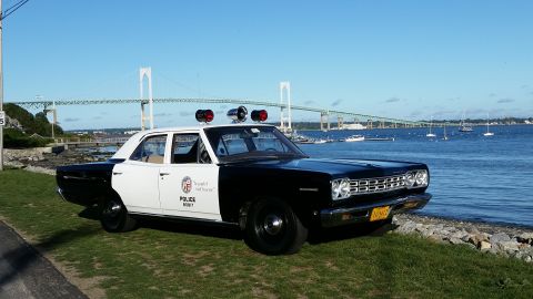 1991 Ford LTD Crown Victoria - A black and white 1968 Plymouth Satellite police car is parked on a grassy area next to a body of water with a bridge in the background.