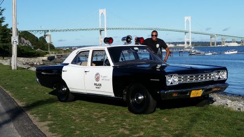 1991 Ford LTD Crown Victoria - A man poses with a vintage black and white police car parked by the water with a bridge in the background.