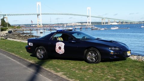 1991 Ford LTD Crown Victoria - A dark blue police car with a light bar and a shield emblem on the door is parked on a grassy area near the water, with a large bridge visi…