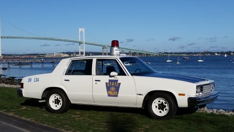 1991 Ford LTD Crown Victoria - A white 1980s Chevrolet Caprice police car is parked near the water, with a bridge and sailboats visible.