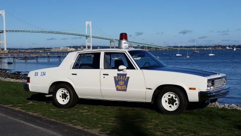 1991 Ford LTD Crown Victoria - A white police car with a black hood and a red light on top is parked by the water with a bridge in the background.