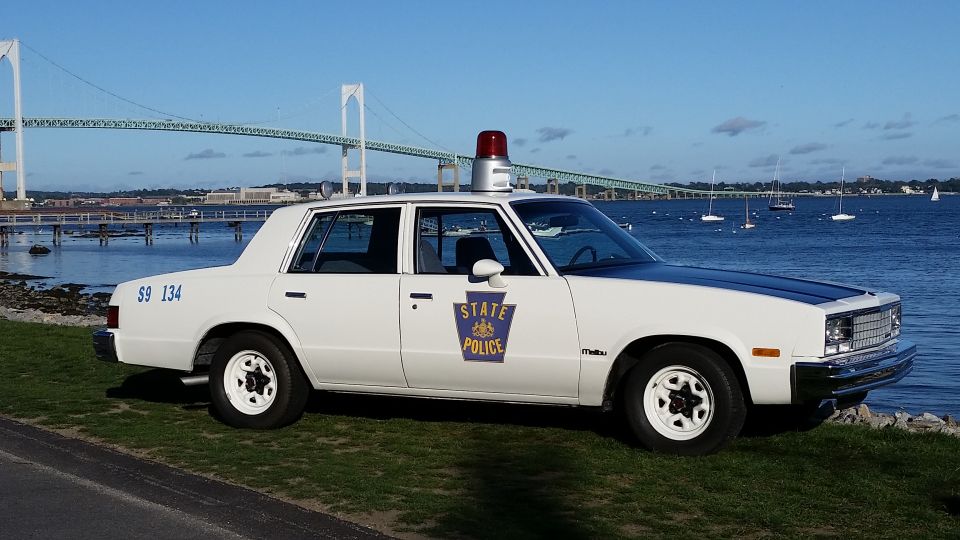 White police car with a black hood and a red light on top, parked on grass by the water