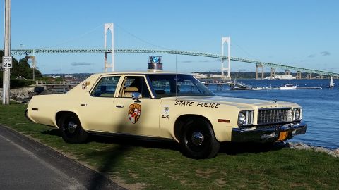 1991 Ford LTD Crown Victoria - A 1977 Plymouth Fury police car is parked by the water with a bridge in the background.