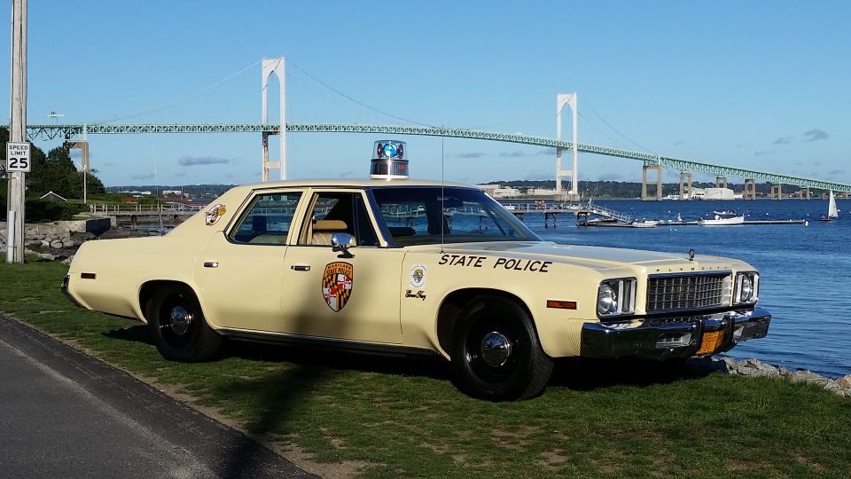Cream-colored 1977 Plymouth Fury police car with 'STATE POLICE' text and a light bar, parked by the water