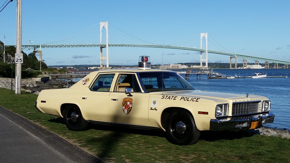 Cream police car with "STATE POLICE" text and Maryland emblem, parked by the water with a bridge in the background