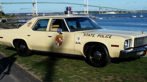 1991 Ford LTD Crown Victoria - This vintage police car, likely a 1977 Plymouth Fury, is parked by the water with a bridge in the background.