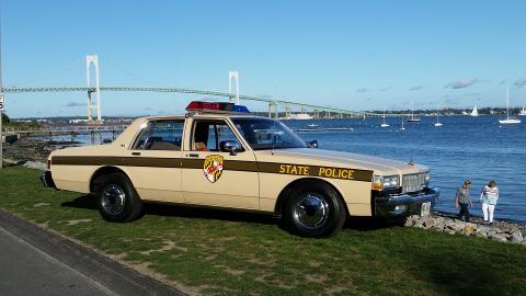 1991 Ford LTD Crown Victoria - A tan police car with a light bar sits by the water, with a bridge and sailboats in the background.