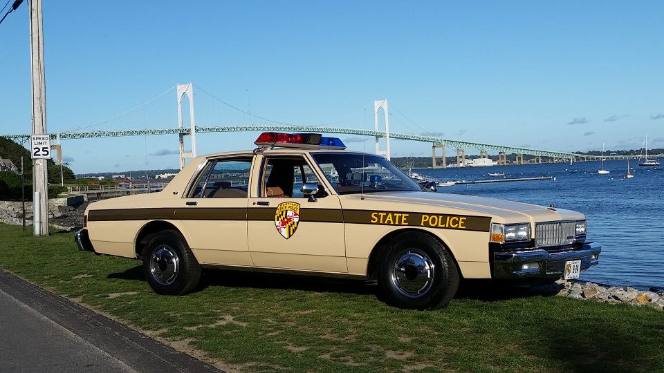 tan police car with "STATE POLICE" on the side, parked by the water with a bridge in the background