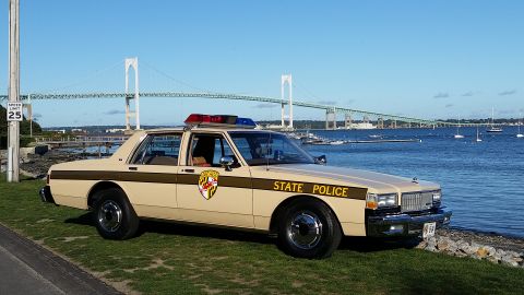 1991 Ford LTD Crown Victoria - A tan 1980s state police car is parked near the water, with a large bridge visible in the distance.