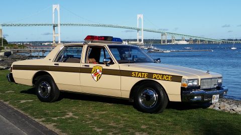 1991 Ford LTD Crown Victoria - A tan police car with "STATE POLICE" text and a Maryland state seal is parked by the water, with a large bridge in the background.