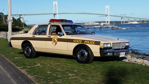 1991 Ford LTD Crown Victoria - A vintage state police car is parked by the water with a large bridge in the background.