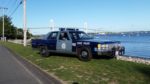1991 Ford LTD Crown Victoria - A blue and gray police car is parked by the water with a bridge in the distance.