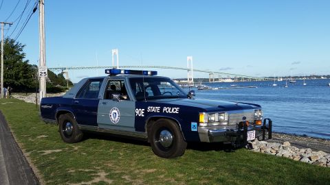 1991 Ford LTD Crown Victoria - This police car is parked on the grass near the water, with a large bridge visible in the distance.