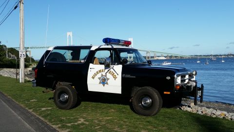 1991 Ford LTD Crown Victoria - A black Dodge Ramcharger marked as "Highway Patrol" sits parked near the water, with a large bridge visible in the distance.