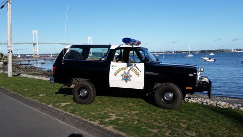 1991 Ford LTD Crown Victoria - A Dodge Ramcharger police vehicle is parked by the water with a bridge in the background.