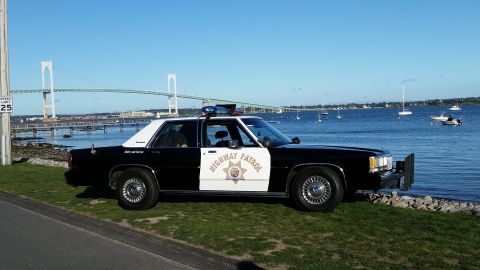 1991 Ford LTD Crown Victoria - A black and white police car is parked on a grassy area next to the water, with a large bridge visible in the distance.