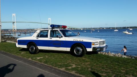 1991 Ford LTD Crown Victoria - A police car is parked by the water with boats and a bridge in the distance.