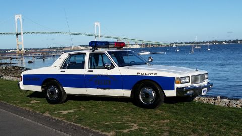 1991 Ford LTD Crown Victoria - A police car is parked on the grass near the water, with a large bridge and sailboats visible in the distance.