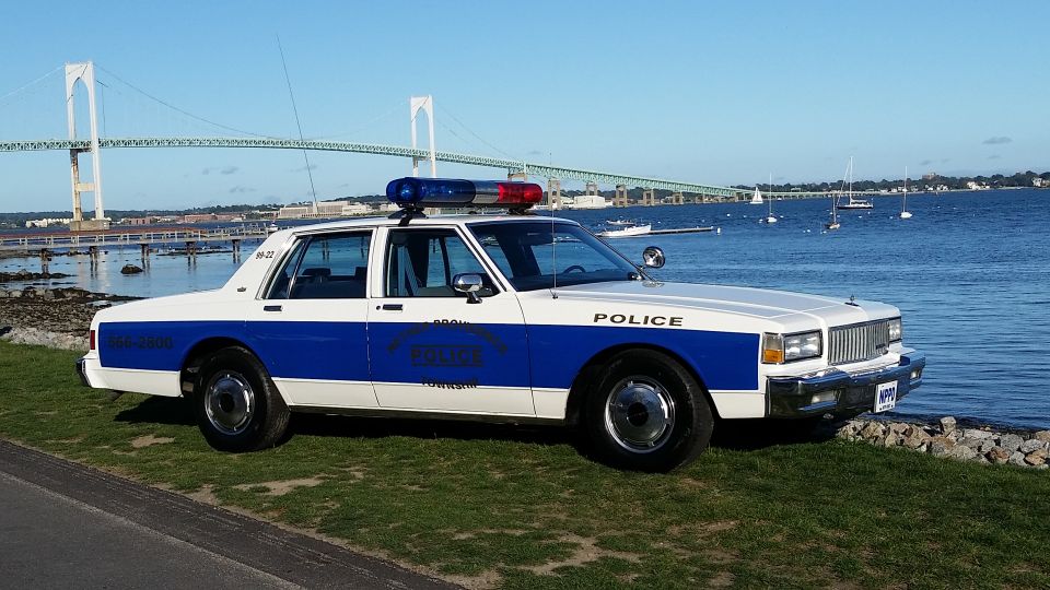 White and blue police car parked by the water, with a bridge in the background