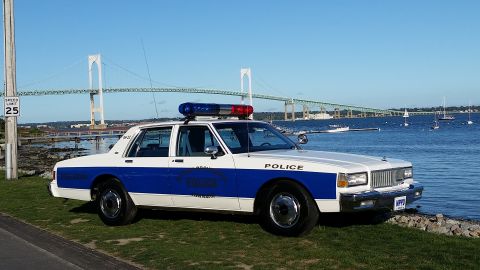 1991 Ford LTD Crown Victoria - Police car with emergency lights parked near a body of water, a bridge spans the horizon.