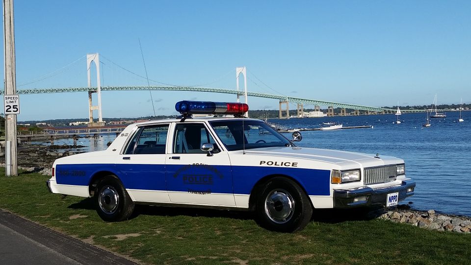 White and blue police car parked on grass by the water, with a large bridge in the background. License plate reads NPPO