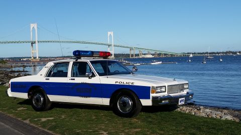 1991 Ford LTD Crown Victoria - A white and blue police car sits by the water with a large bridge spanning the bay in the distance.