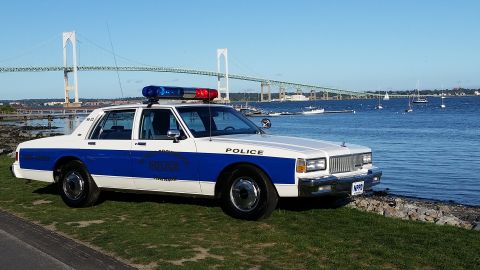 1991 Ford LTD Crown Victoria - A white and blue police car is parked near the water with a bridge in the distance.