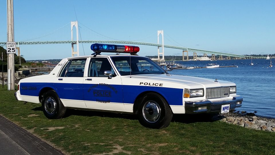 Police car with blue and white livery parked on grass near water. Bridge and boats in background