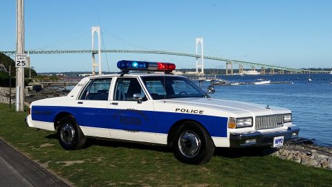 1991 Ford LTD Crown Victoria - A white and blue police car is parked on a grassy area next to the water, with a large bridge in the distance.