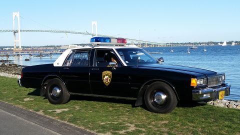 1991 Ford LTD Crown Victoria - This police car, likely from the 1980s, is parked by the water with a bridge in the background.
