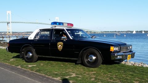1991 Ford LTD Crown Victoria - Side view of a vintage police car parked by the water with a bridge and sailboats in the distance.