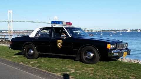 1991 Ford LTD Crown Victoria - A black police car with a light bar is parked on the grass near the water, with a bridge and sailboats visible.