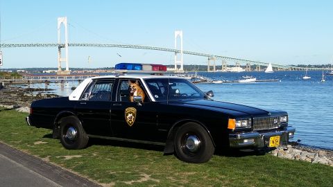 1991 Ford LTD Crown Victoria - Photograph of a vintage black-and-white police sedan parked on grass by a waterfront with a suspension bridge in the background.