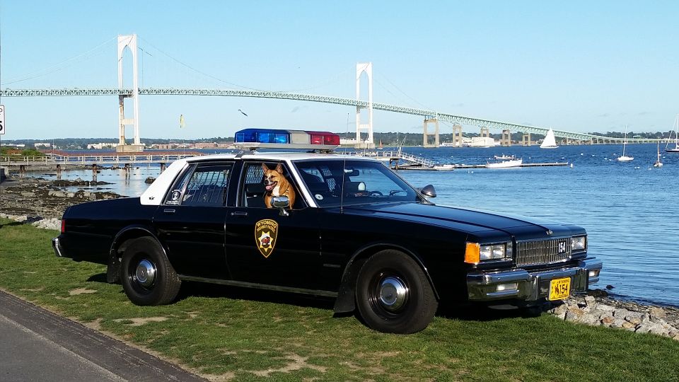 Black and white classic police sedan parked by a harbor with a suspension bridge behind