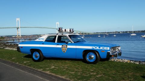1991 Ford LTD Crown Victoria - A vintage police car sits by the water, with a bridge and sailboats visible in the distance under a clear blue sky.