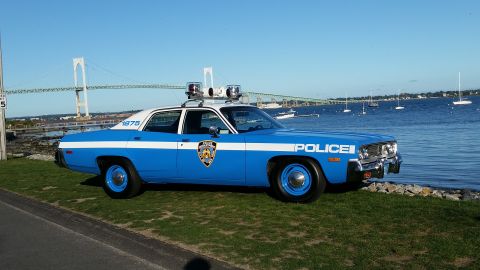 1991 Ford LTD Crown Victoria - A blue and white police car with "1975" on its side is parked on the grass near a body of water with a bridge in the background.
