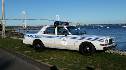 1991 Ford LTD Crown Victoria - Photograph of a white vintage police sedan with blue striping parked near a waterfront with a suspension bridge.