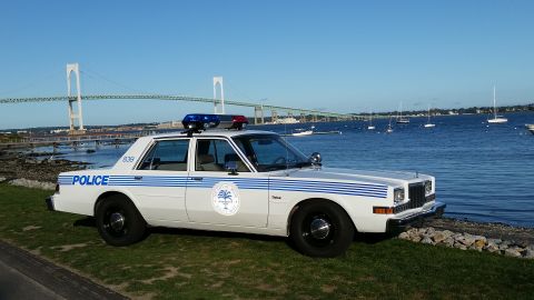1991 Ford LTD Crown Victoria - Photograph of a white police sedan with blue striping and roof lights parked at a harbor with a bridge behind.
