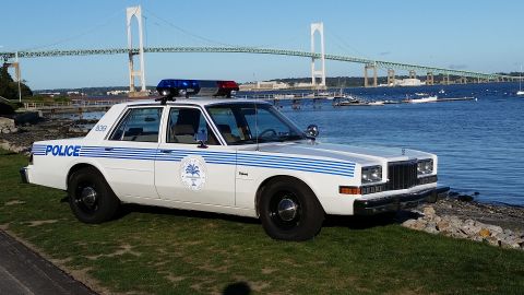 1991 Ford LTD Crown Victoria - Photograph of a vintage white-and-blue police sedan parked by a harbor with a suspension bridge in the background.