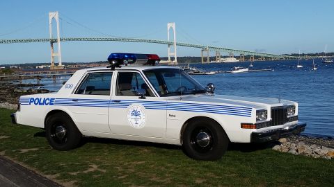 1991 Ford LTD Crown Victoria - Photograph of a white police sedan with blue striping parked near a waterfront with a suspension bridge.