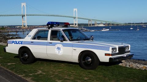 1991 Ford LTD Crown Victoria - Photograph of a white police sedan with blue striping parked beside a harbor and suspension bridge.